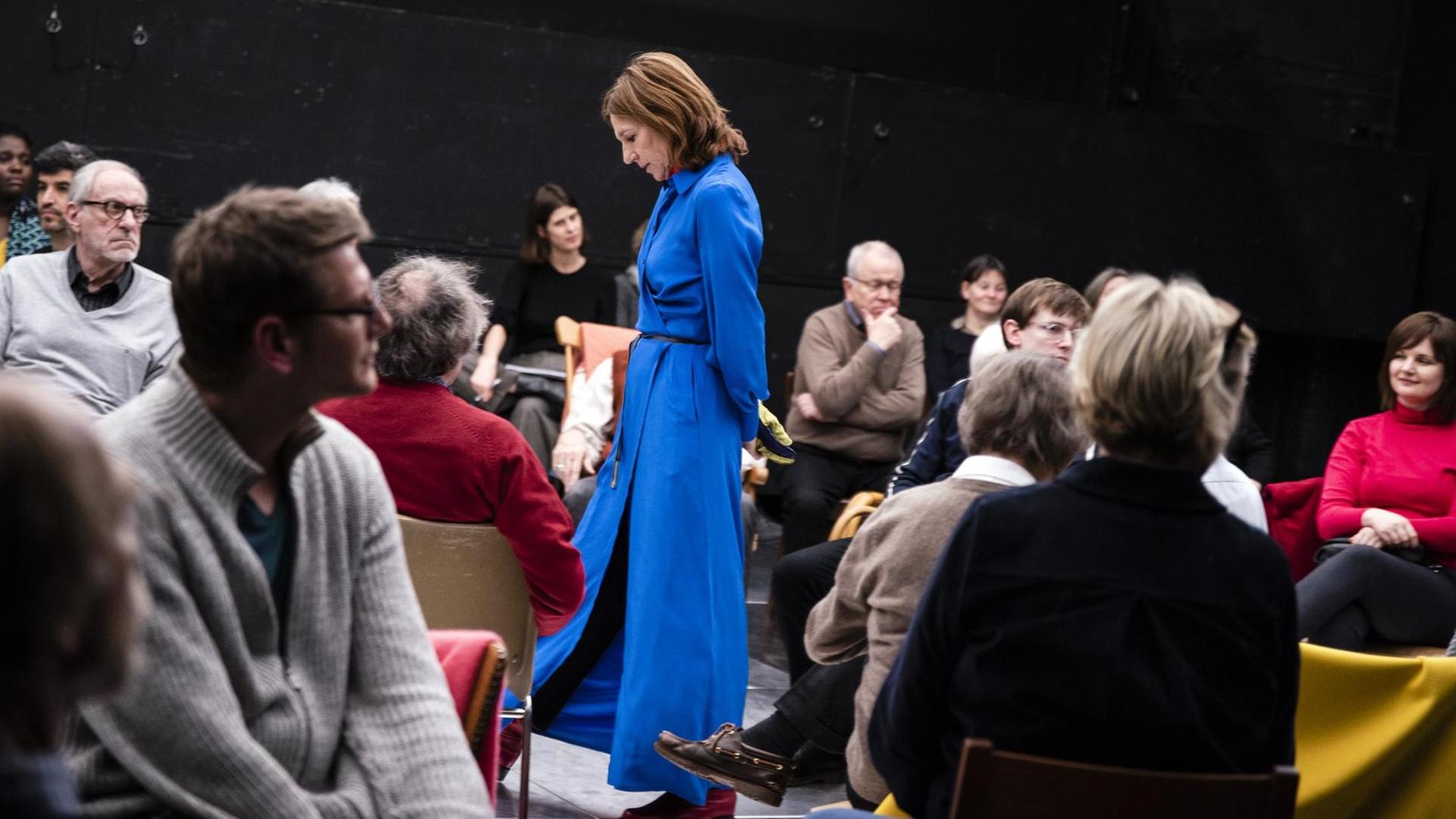 Die Schauspielerin Elsie de Brauw steht in einen langen blauen Mantel gekleidet in den Zuschauerreihen im Schauspielhaus Bochum. Ihr Kopf ist gesenkt.