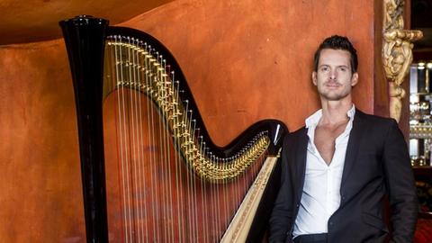 Xavier de Maistre lehnt mit seiner Harfe an einer Wand, im Hintergrund Bar mit Tangotänzerin im roten Kleid