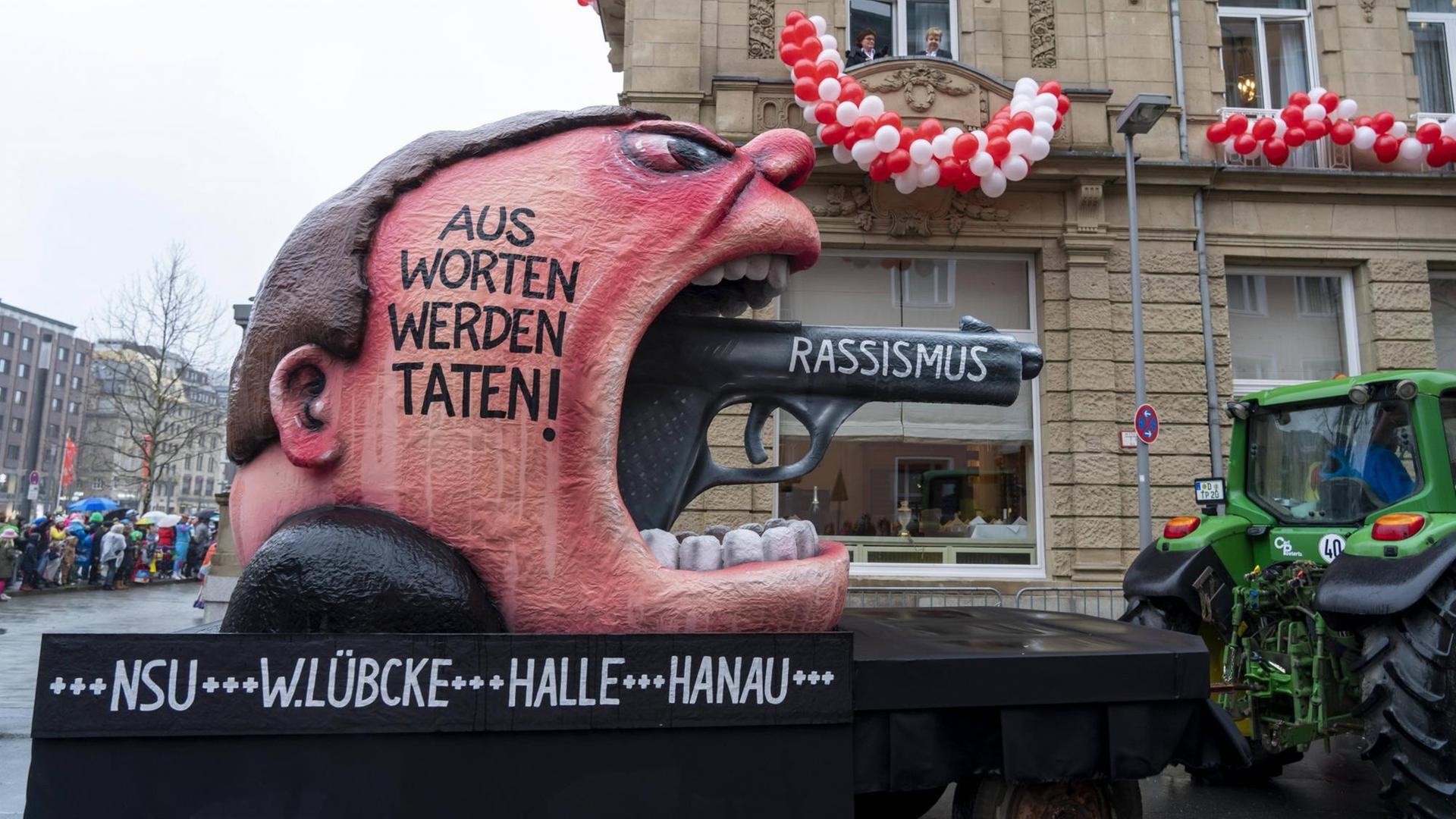 Ein Karnevalswagen mit überdimensionalem Kopf aus dessen Mund eine Pistole ragt. 