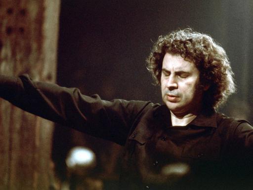 Mikis Theodorakis steht mit ausgebreiteten Armen als Dirigent vor einem Orchester.