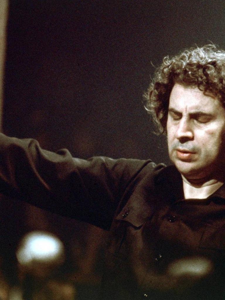 Mikis Theodorakis steht mit ausgebreiteten Armen als Dirigent vor einem Orchester.