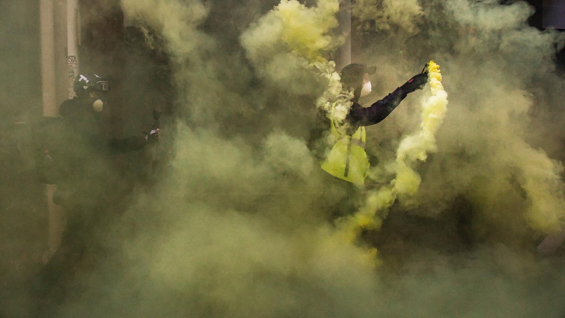 Zwei Männer, einer in einer gelben Warnweste, stehen im gelblichen Rauch von gezündeter Pyrotechnik.