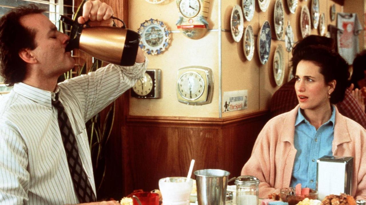 Der Meteorologe Phil, gespielt von Bill Murray, sitzt neben seiner Aufnahmeleiterin Rita Hanson, gespielt von Andie MacDowell, an einem Tisch und trinkt Kaffee direkt aus einer Kaffeekanne.