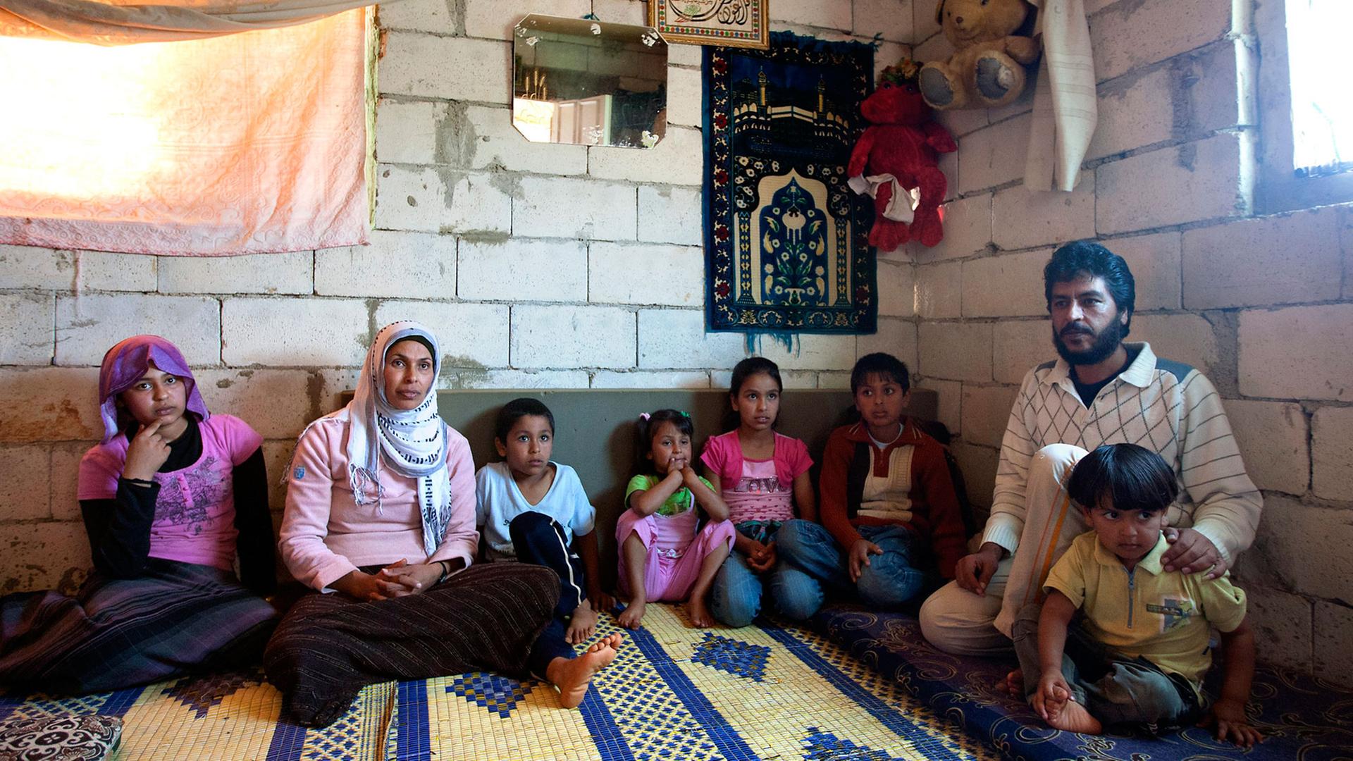 Eine syrische Familie - Mutter, Vater und sechs Kinder - sitzt in einer Wohnung mit unverputzten Wänden auf einem Teppich, an den Wänden hängen ein kleiner Spiegel, einzelne Bilder und zwei große Stoff-Teddybären.