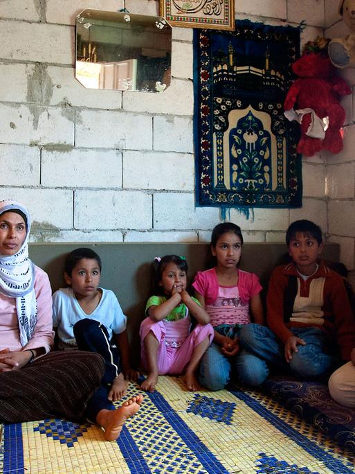 Eine syrische Familie - Mutter, Vater und sechs Kinder - sitzt in einer Wohnung mit unverputzten Wänden auf einem Teppich, an den Wänden hängen ein kleiner Spiegel, einzelne Bilder und zwei große Stoff-Teddybären.