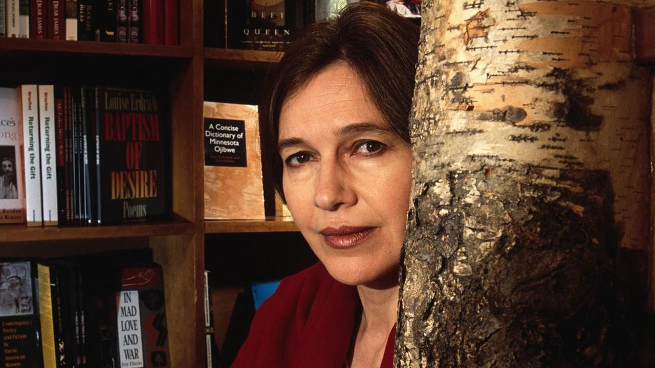 Die Schriftstellerin Louise Erdrich blickt in die Kamera, hinter ihr sieht man ein Bücherregal und rechts neben ihr einen Baumstamm.