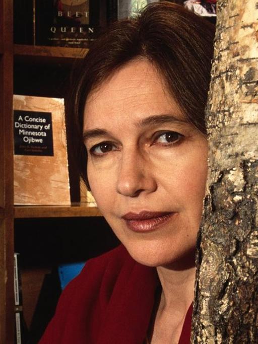 Die Schriftstellerin Louise Erdrich blickt in die Kamera, hinter ihr sieht man ein Bücherregal und rechts neben ihr einen Baumstamm. Die Schriftstellerin Louise Erdrich blickt in die Kamera, hinter ihr sieht man ein Bücherregal und rechts neben ihr einen Baumstamm.