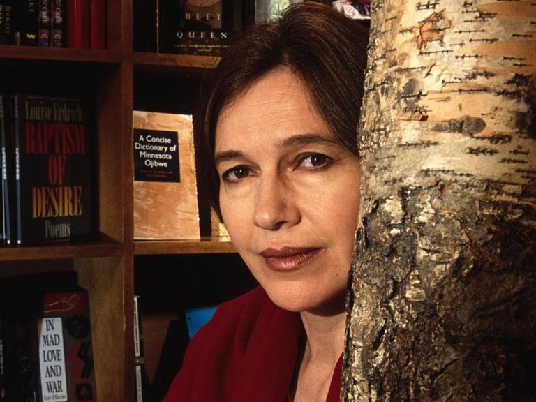 Die Schriftstellerin Louise Erdrich blickt in die Kamera, hinter ihr sieht man ein Bücherregal und rechts neben ihr einen Baumstamm. Die Schriftstellerin Louise Erdrich blickt in die Kamera, hinter ihr sieht man ein Bücherregal und rechts neben ihr einen Baumstamm.