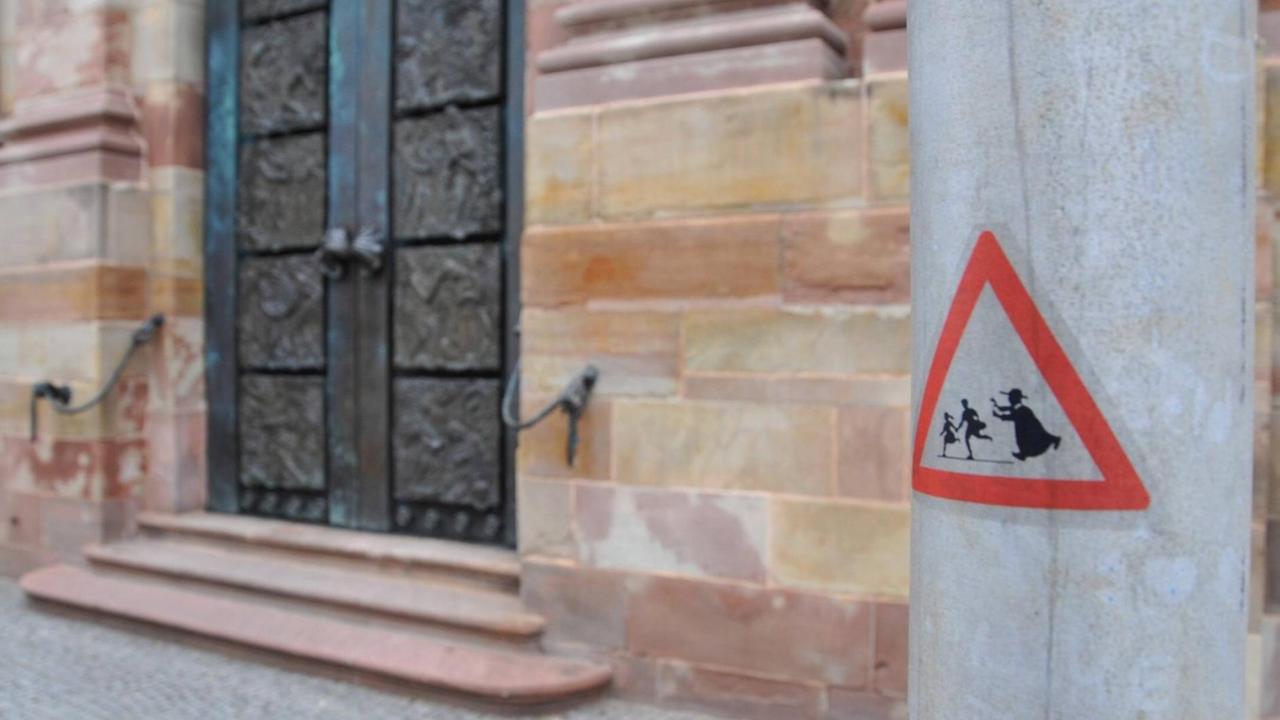 Vor einer Kirchetür im Hintergrund klebt auf einem Pfahl im Vordergrund ein Warnschild. Darauf ein geistlicher, der zwei Kindern hinterher läuft.