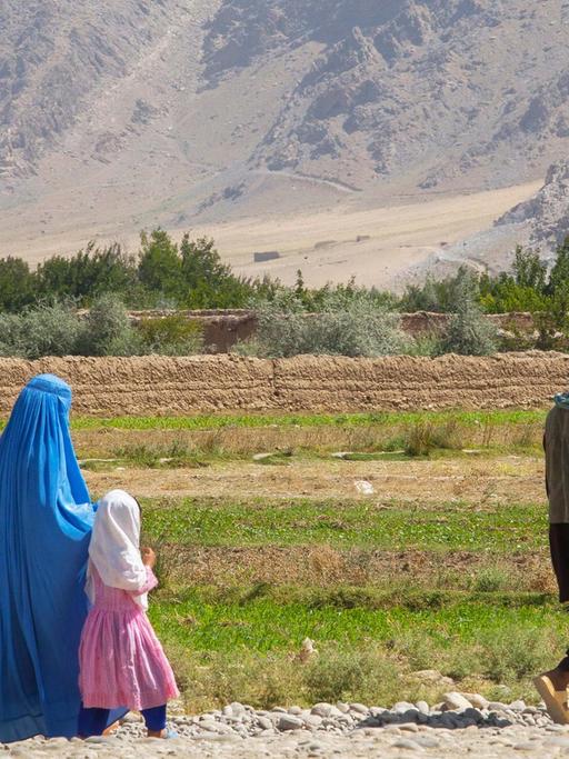 Eine afghanische Familie spaziert über eine Geröllstraße in der Region Kapisa. Der Mann läuft zwei Meter vorneweg, Mutter und Tochter, beider verschleiert, hinterher. Im Hintergrund Ackerflächen und hohes Gebirge.