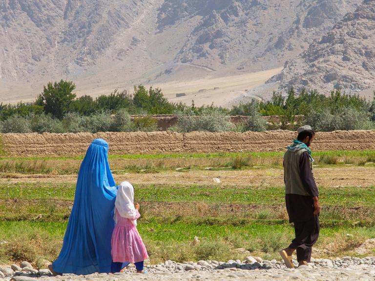 Eine afghanische Familie spaziert über eine Geröllstraße in der Region Kapisa. Der Mann läuft zwei Meter vorneweg, Mutter und Tochter, beider verschleiert, hinterher. Im Hintergrund Ackerflächen und hohes Gebirge.