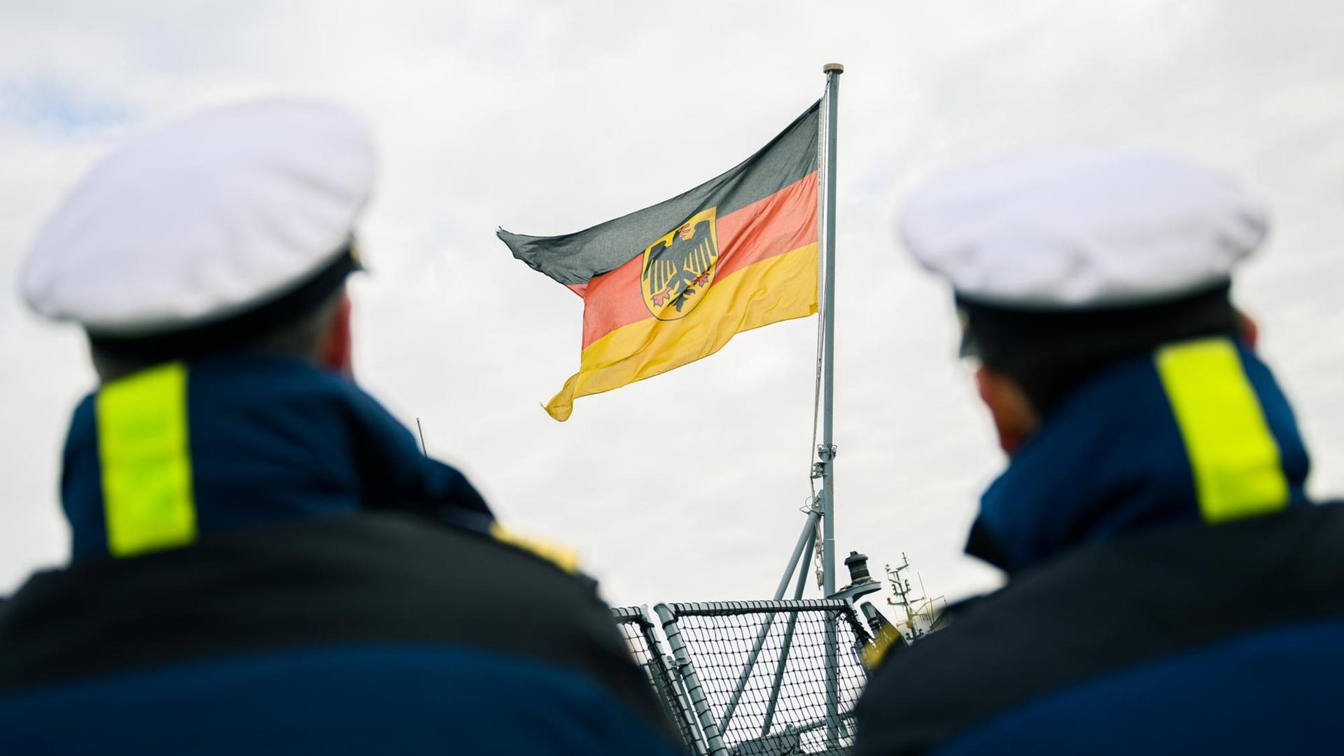 Zwei Marinesoldaten stehen am Hafen des Marinestützpunktes Wilhelmshaven, zwischen ihnen ist die deutsche Flagge, die auf dem Heck der Fregatte angebracht ist, zu sehen.