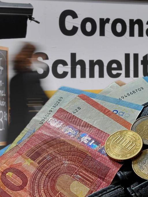 Eine Geldbörse voller Scheine vor einem Corona-Schnelltest-Schild