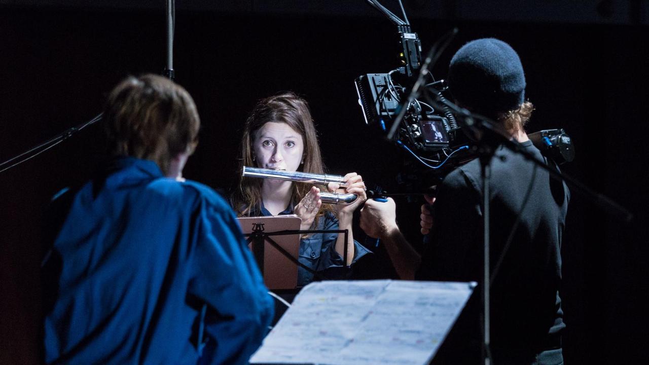 Eine junge Frau vor schwarzem Hintergrund bläst in eine Flöte, dicht vor ihr stehen rückwärtig ein Kameramann und eine Person im blauen Oberteil.