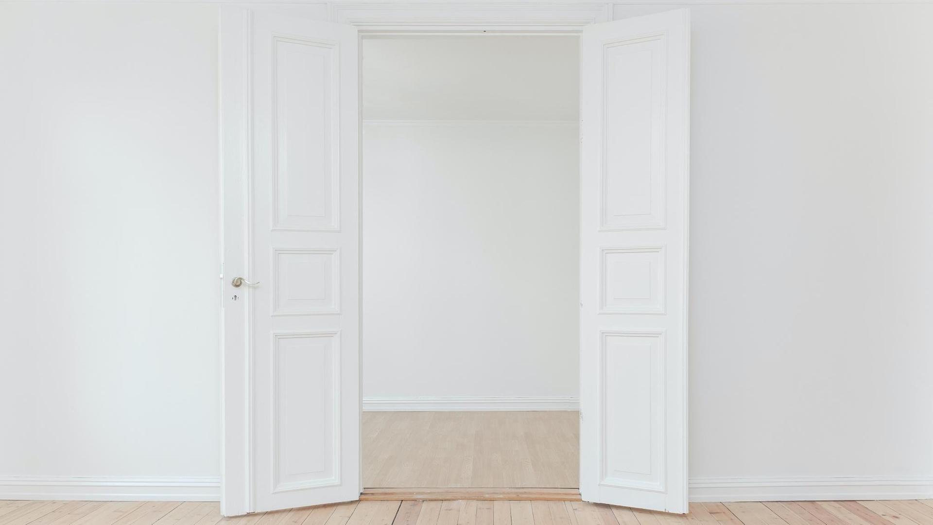 Eine offene Tür in einem leeren Altbau mit Holzfussboden und weißen Wänden.
