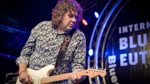 Ein lockiger Mann in buntem Hemd steht auf einer Bühne und spielt E-Gitarre