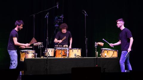 Drei junge Schlagzeuger auf einer Bühne mit verschiedenen Schlagwerken in Aktion.