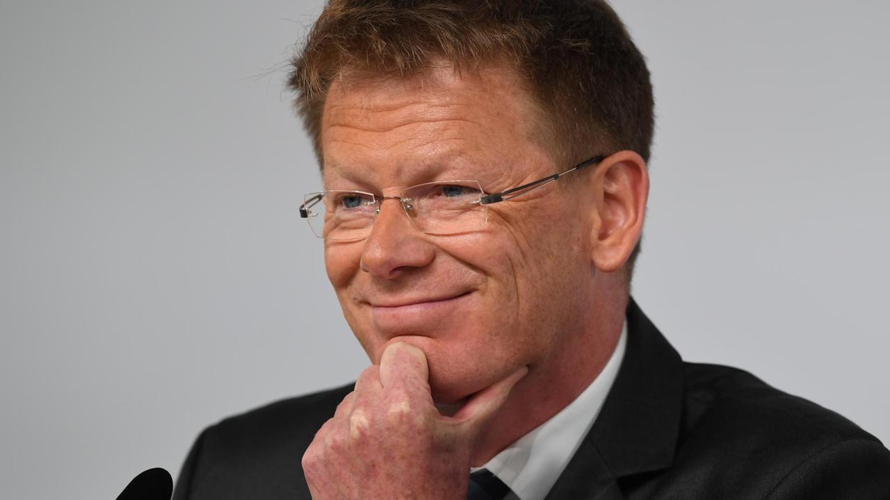 Lutz sitzt vor einem Mikrofon, lächelt und hat eine Hand am Kinn. Er trägt eine Brille.
