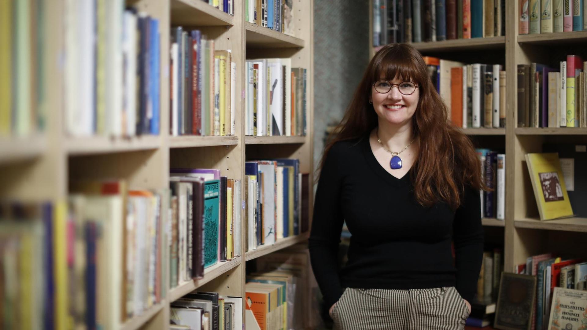 Allison Devers steht in ihrem Buchladen "Second Shelf" in London, umgeben von gefüllten Bücherregalen und lächelt in die Kamera, die Hände in den Hosentaschen.