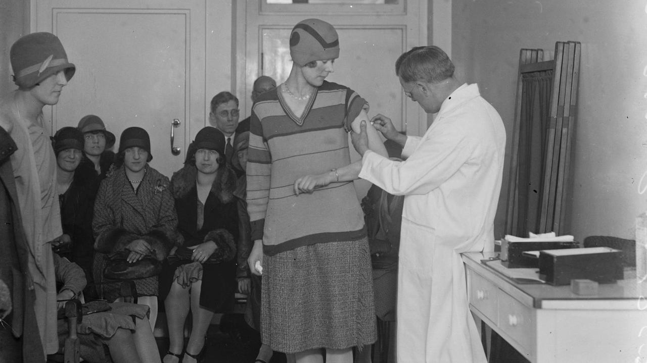 Pockenimpfung im April 1929 in London. Ein Arzt impft in einem Arztzimmer eine Frau. Im Hintergrund warten Frauen und Männer.