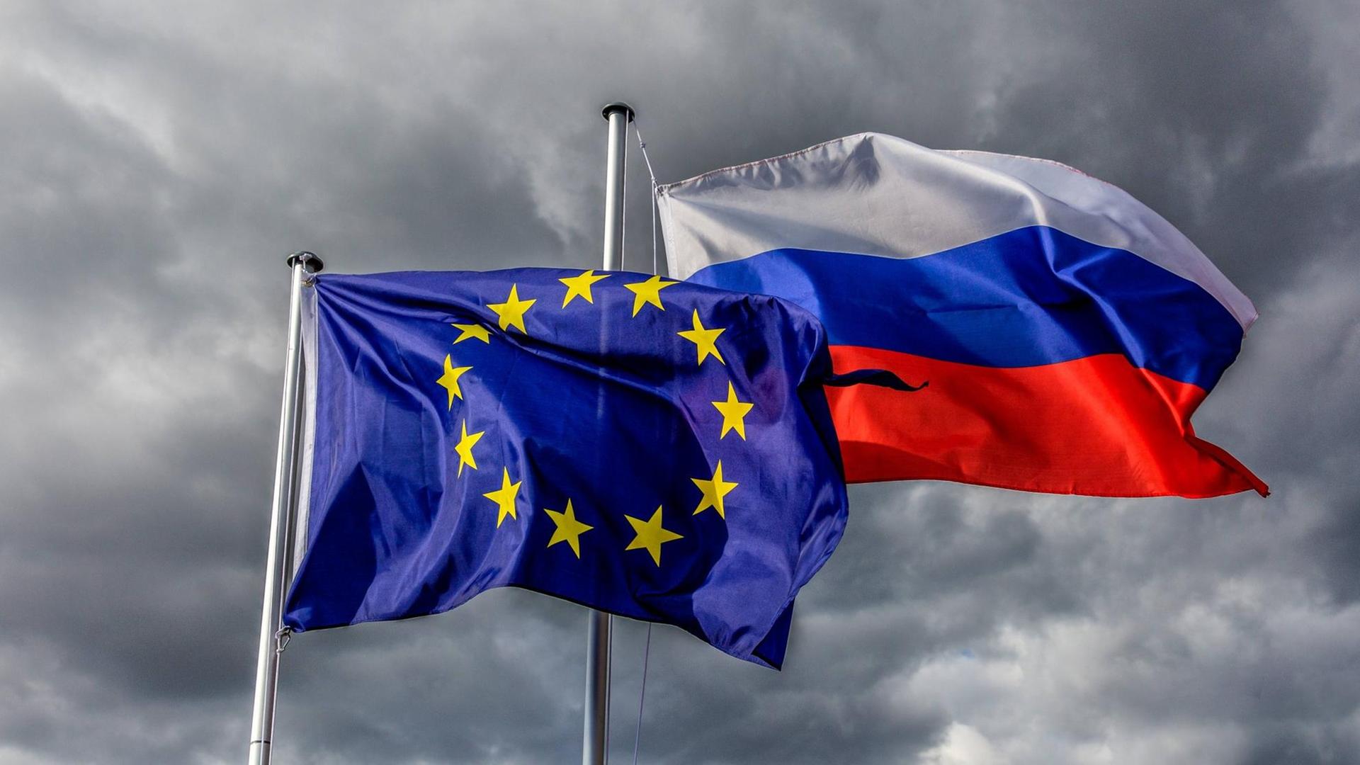 Europaflagge und Flagge der Russischen Föderation wehen vor grauem Himmel im Wind.