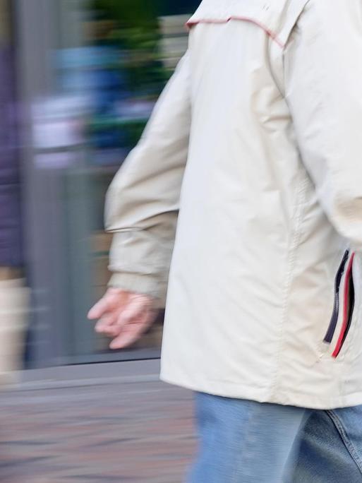 Ein älterer Passant geht durch eine Fußgängerzone und hält eine Mundschutzmaske in der Hand. Im Hintergrund geht weiterer Passant, der einen Mundschutz trägt.