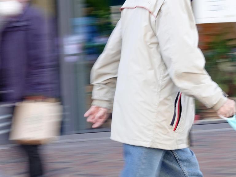 Ein älterer Passant geht durch eine Fußgängerzone und hält eine Mundschutzmaske in der Hand. Im Hintergrund geht weiterer Passant, der einen Mundschutz trägt.