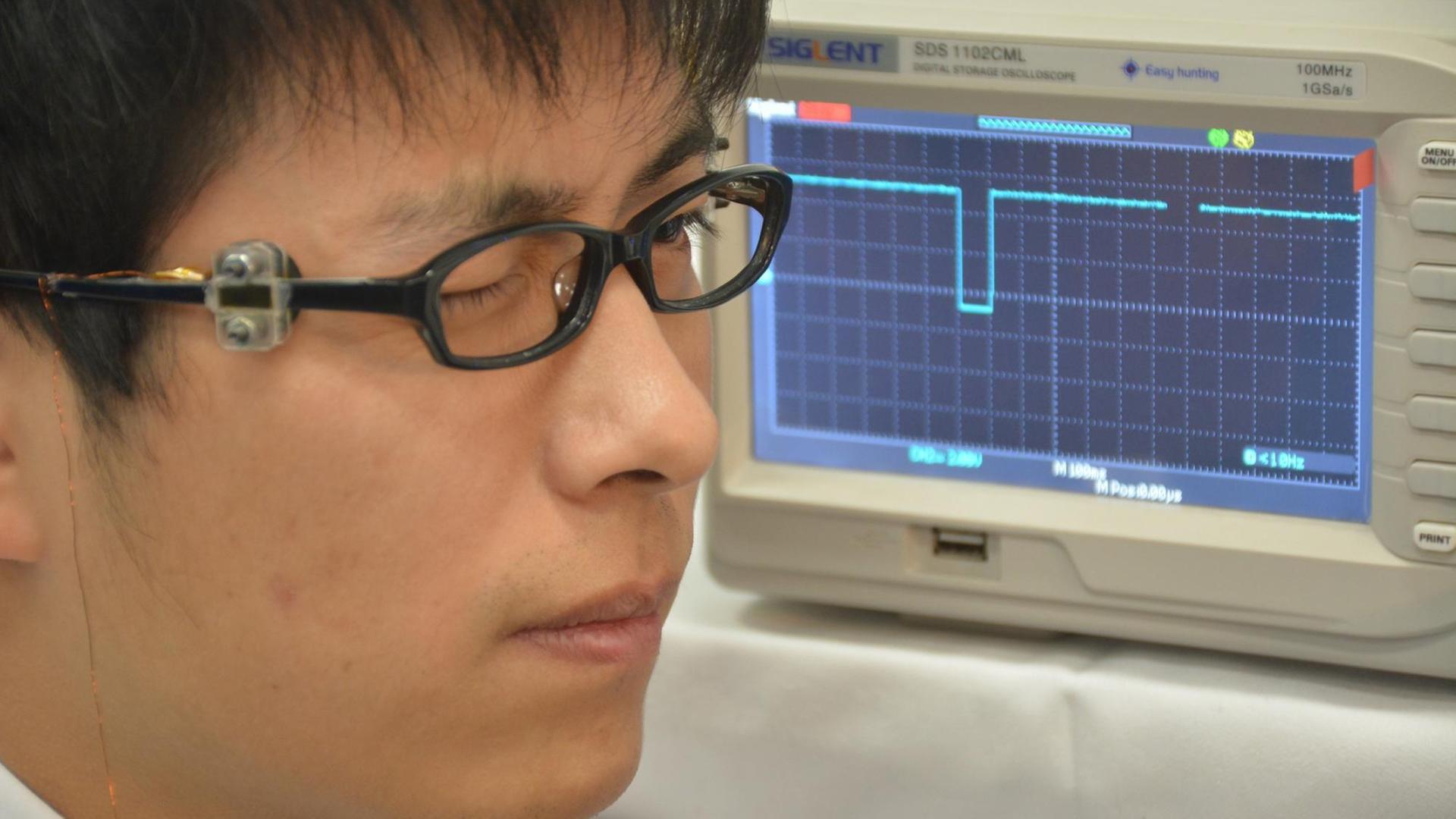 Ein Mann trägt eine Brille mit einem Sensor und blinzelt. Im Hintergrund sieht man einen Computerbildschirm, der einen Graphen zeigt.