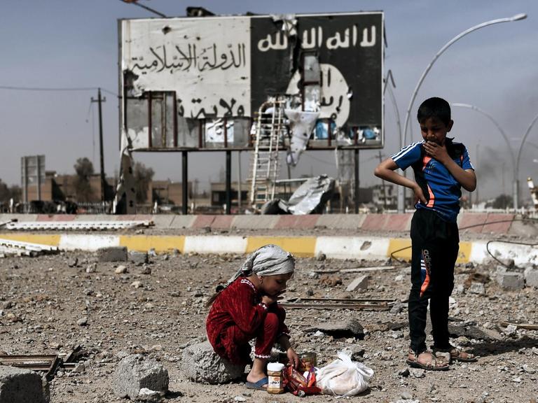 Iraqi children sit amidst the rubble of a street in Mosul's Nablus neighbourhood infront of a billboard bearing the logo of the Islamic State (IS) group on March 12, 2017, during an offensive by security forces to retake the western parts of the city from IS fighters. / AFP PHOTO / ARIS MESSINIS