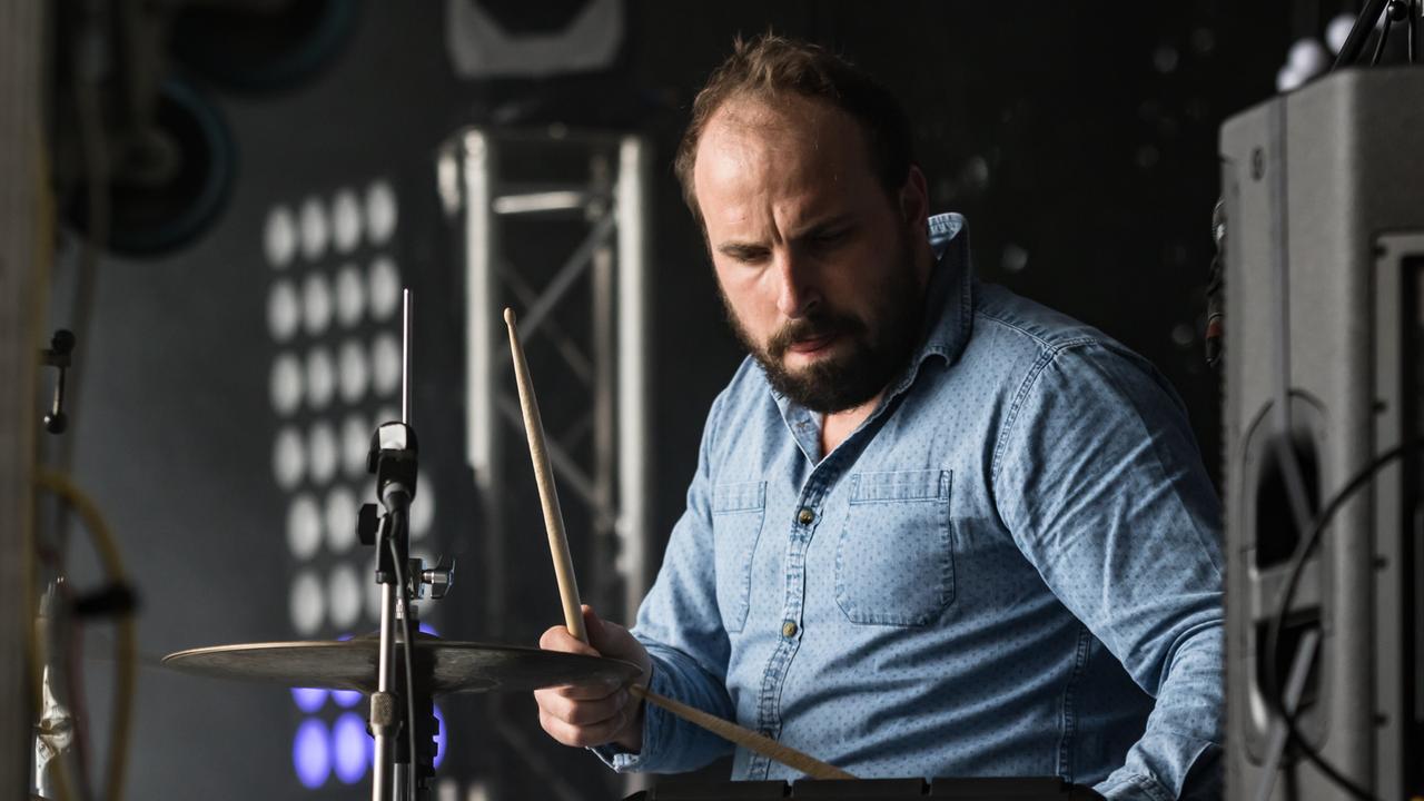 Ein Mann in einem hellblauen Hemd sitz an einem Schlagzeug und spielt. 
