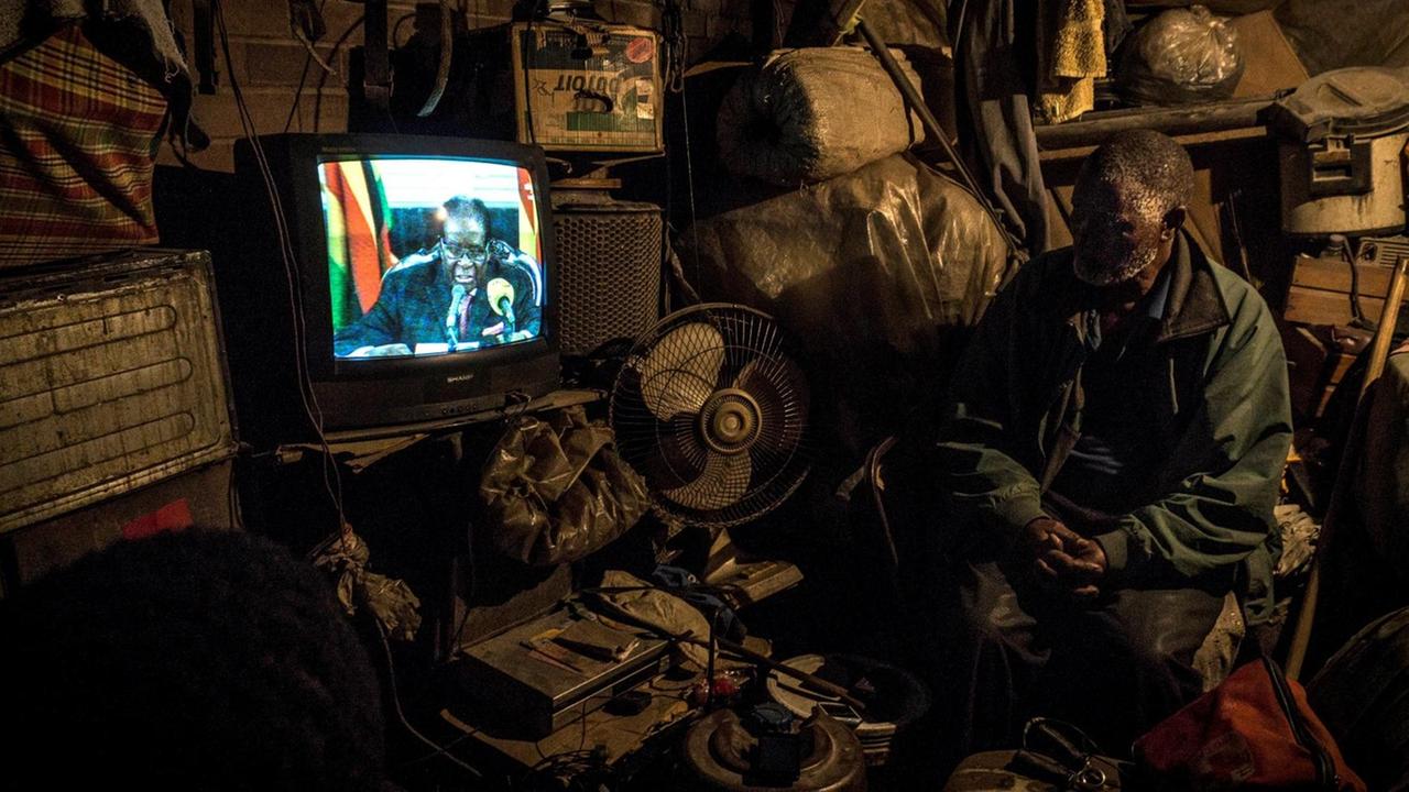 Es ist düster in der Hütte: Der ältere Mann mit Bart sitzt auf einem Stuhl und verfolgt auf einem alten Fernseher Mugabes Ansprache. Die Hütte ist mit allerlei Schrott und Krimskrams vollgestopft.