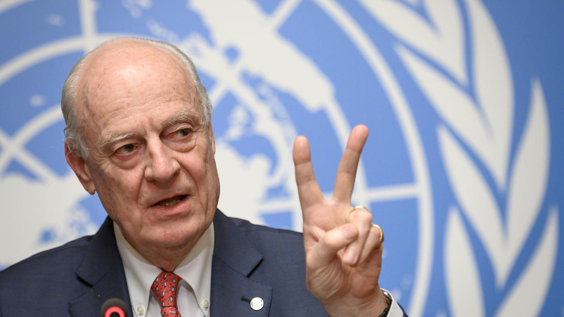 Uno-Syrienbeauftragter Staffan de Mistura bei der Presskonferenz zur Einigung auf einen Verfassungsauschusses für Syrien in Genf. Er steht vor einer blauen Wand mit dem Wappen der Vereinten Nationen und formt sein linke Hand zum Peace-Zeichen, indem er Zeige- und Mittelfinger hebt.