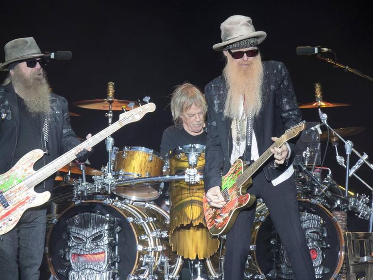 Zwei Männer mit langem Bart, Sonnenbrille und Hut stehen auf einer Bühne. Der linke spielt E-Bass, der rechte E-Gitarre. Im Hintergrund sitzt ein Schlagzeuger hinter seinem Instrument.