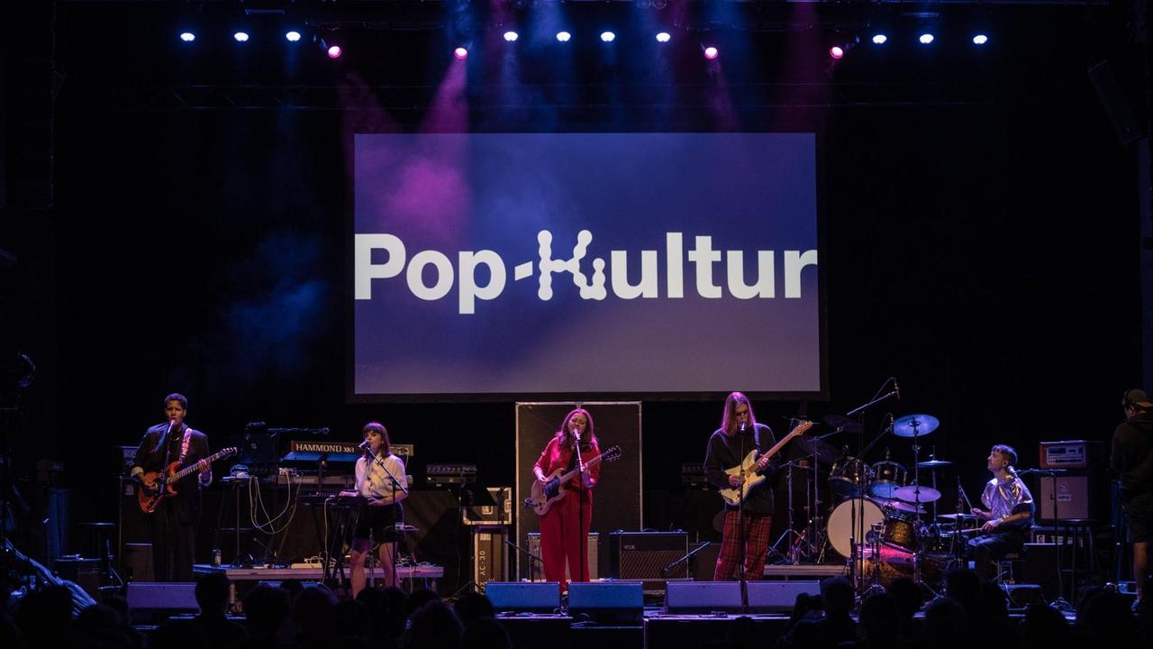Musiker und Musikerinnen stehen auf einer lila erleuchteten Bühne, auf der der Schriftzug Pop-Kultur zu lesen ist.