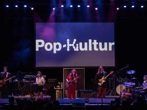 Musiker und Musikerinnen stehen auf einer lila erleuchteten Bühne, auf der der Schriftzug Pop-Kultur zu lesen ist.