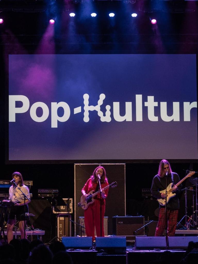 Musiker und Musikerinnen stehen auf einer lila erleuchteten Bühne, auf der der Schriftzug Pop-Kultur zu lesen ist.