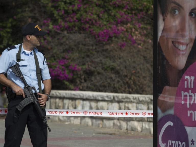 Schwerbewaffneter Polizist steht neben einer Litfaßsäule, auf der eine Frau auf einem Werbeplakat lächelt
