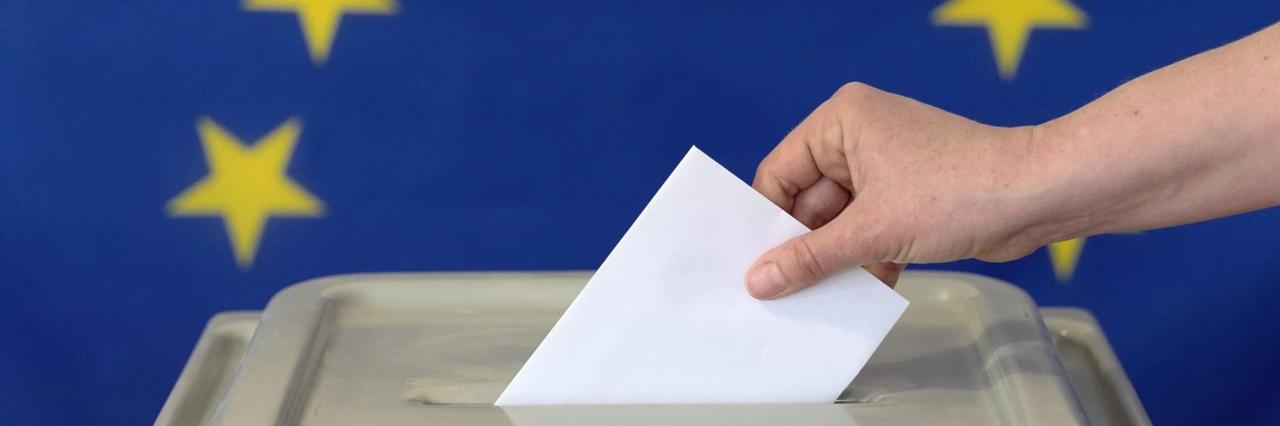 Eine Hand steckt einen Umschlag in eine Wahlurne vor der Europafahne. Symbolfoto.