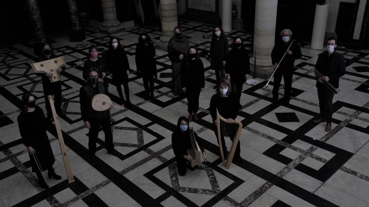 Die Musikerinnen und Musiker des Ensembles Oltremontano stehen im Halbkreis auf einem ornamentierten Steinboden, sie tragen schwarze Konzertkleidung und haben ihre Instrumente bei sich. Sie schauen nach oben in die Kamera und tragen Mundschutz.