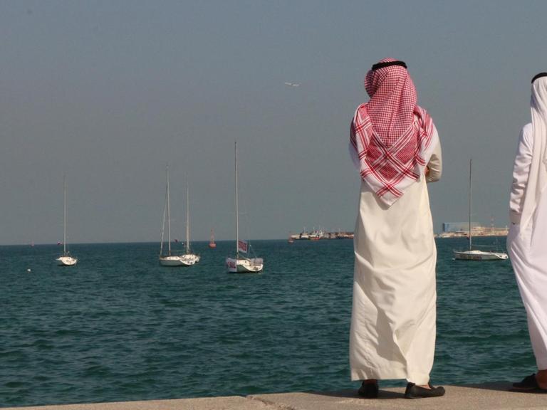 Zwei traditionell gekleidete katarische Bürger stehen an der Uferpromenade der Hauptstadt Doha und blicken auf den Persischen Golf.