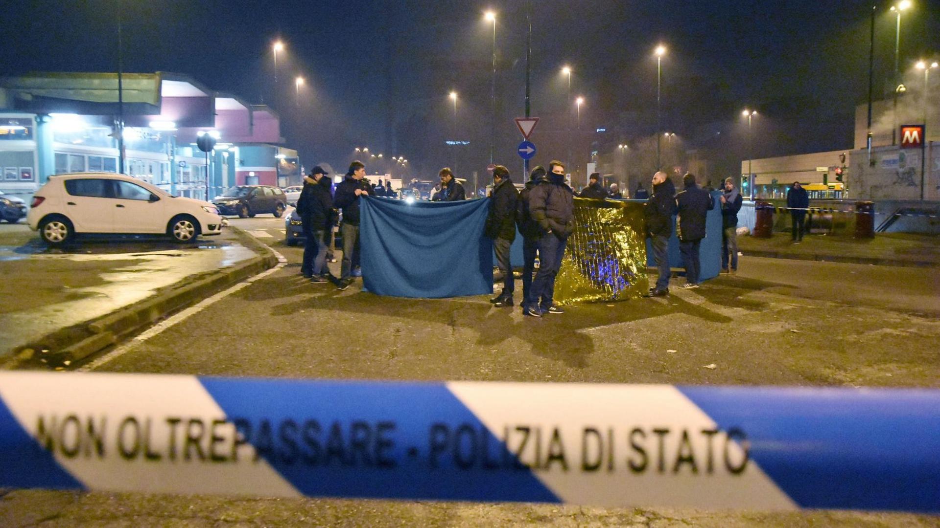 Sie sehen Polizisten in dem Land Italien. Vorne im Bild sehen Sie ein blau-weißes Absperr-Band.