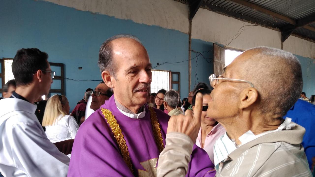 Ein ältere Priester im lila Ornat spricht mit einem älteren Mann im Gottesdienstraum.