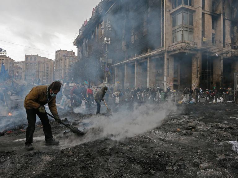 Auf dem verwüsteten Unabhängigkeitsplatz in Kiew versuchen mehrere Männer mit Schaufeln, qualmenden Schutt zu entfernen.