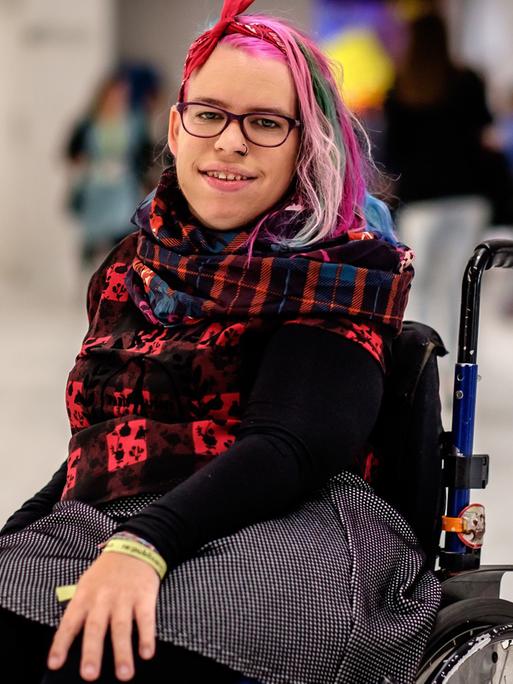 Eine junge Frau mit rosa, grüner und violetter Haarsträhne sitzt in einem Rollstuhl und schaut in die Kamera.
