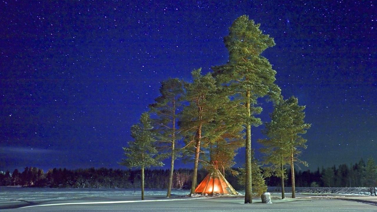 Ein von innen durch Feuer beleuchtetes Zelt unter Bäumen in einer Schneelandschaft in Lappland