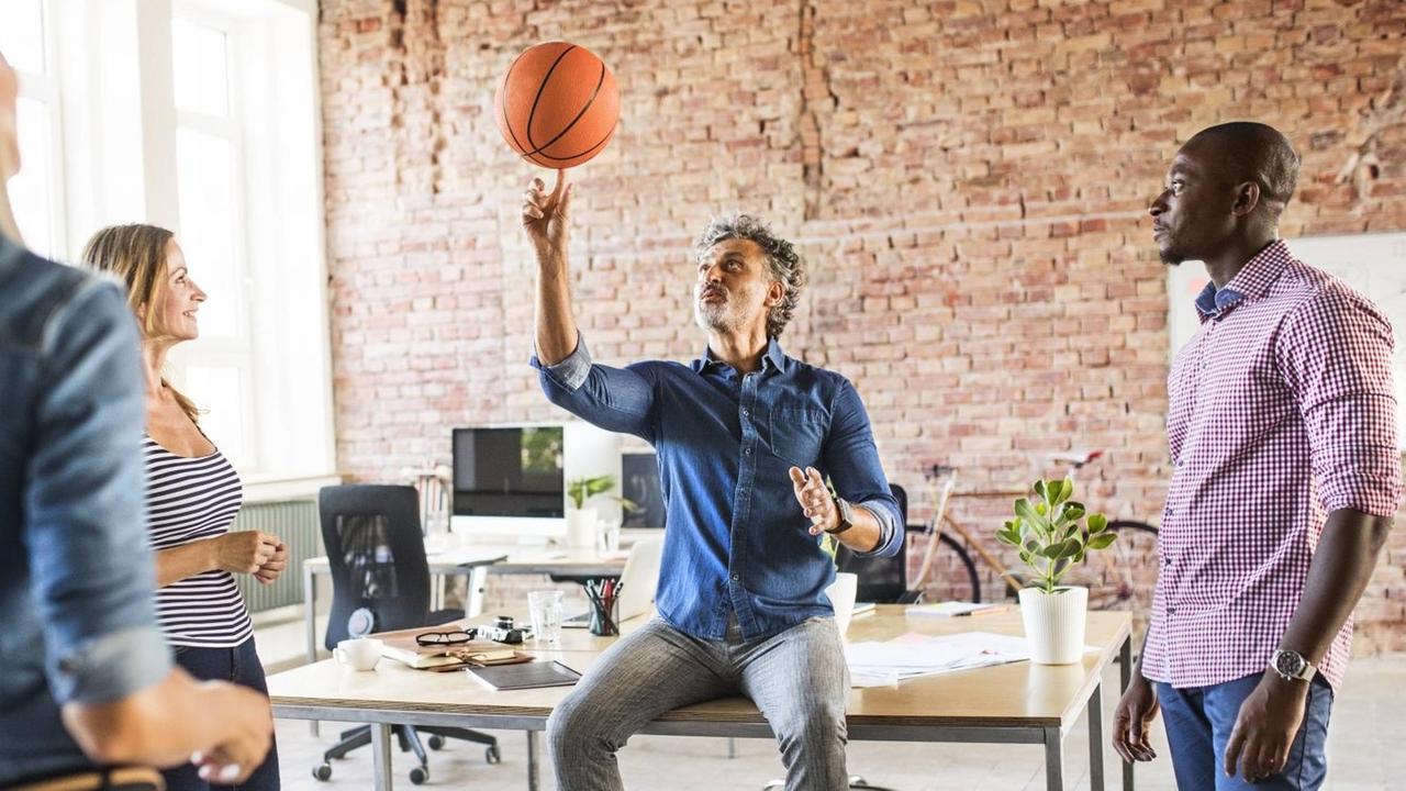 Ein Mann sitzt auf einem Schreibtisch und balanciert einen Basketball auf dem Finger. Um ihn herum stehen Kollegen. Das Büro ist licht, stilvoll und geräumig eingerichtet.