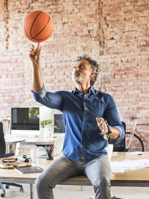 Ein Mann sitzt auf einem Schreibtisch und balanciert einen Basketball auf dem Finger. Um ihn herum stehen Kollegen. Das Büro ist licht, stilvoll und geräumig eingerichtet.