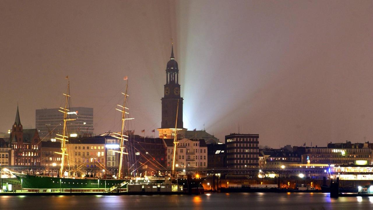 Der Hamburger Hafen erstrahlt in der Nachtbeleuchtung, im Hintergrund ragt der Turm des Kirche laserstrahlend hervor.
