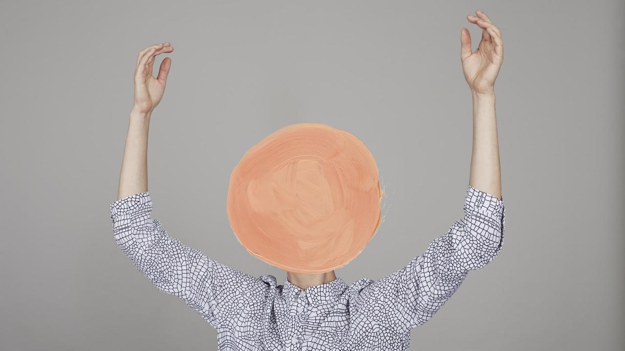 Ein Mensch in gemustertem weißem Hemd hebt die Arme empor – der Kopf ist mit einem orangene Kreis übermalt.