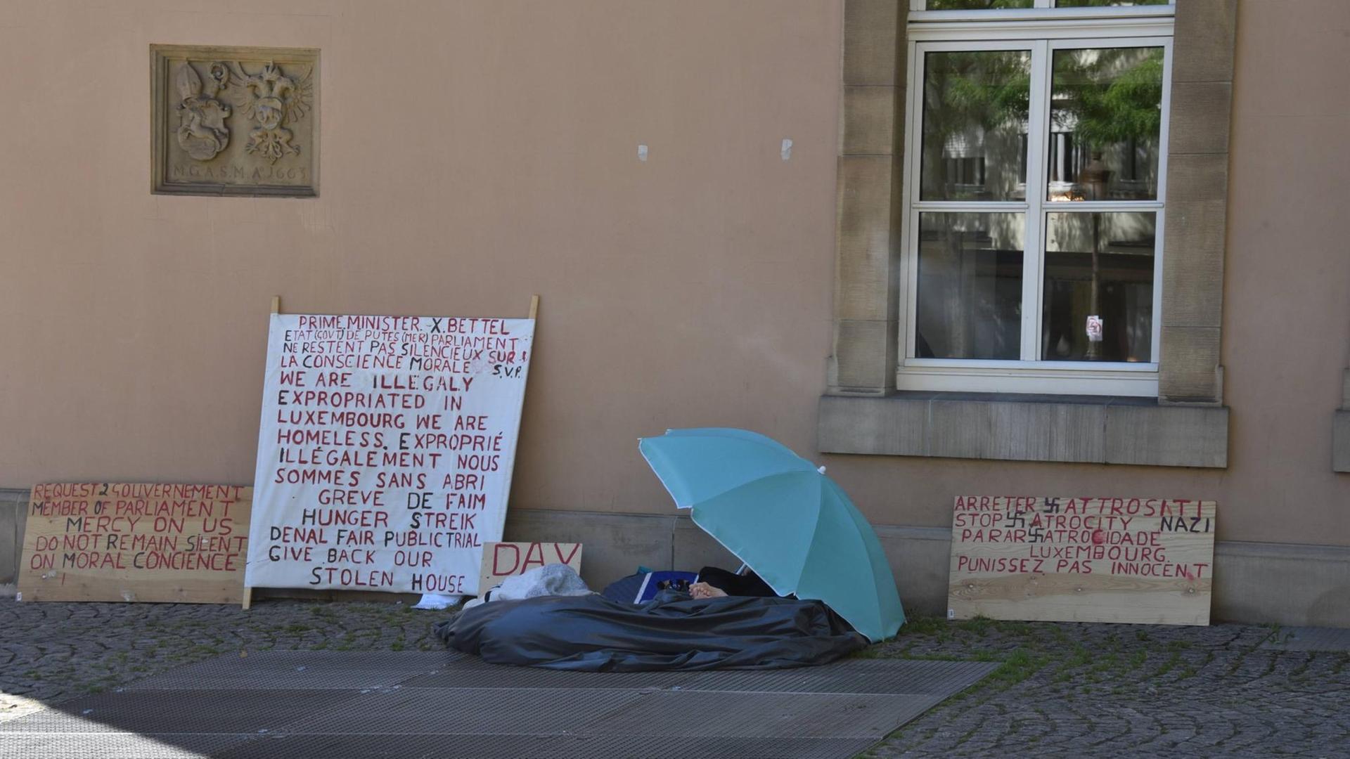 Ein Obdachloser mit Protest-Schildern, hinter einem Regenschirm verborgen, sitzt an einer Hauswand in Luxemburg am Place de Clairefontaine, im August 2016.